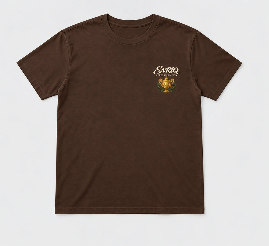 ENRIQ Apres Champion Tee Cocoa – flatlay met gouden trofee-borstlogo (AI-visualisatie)